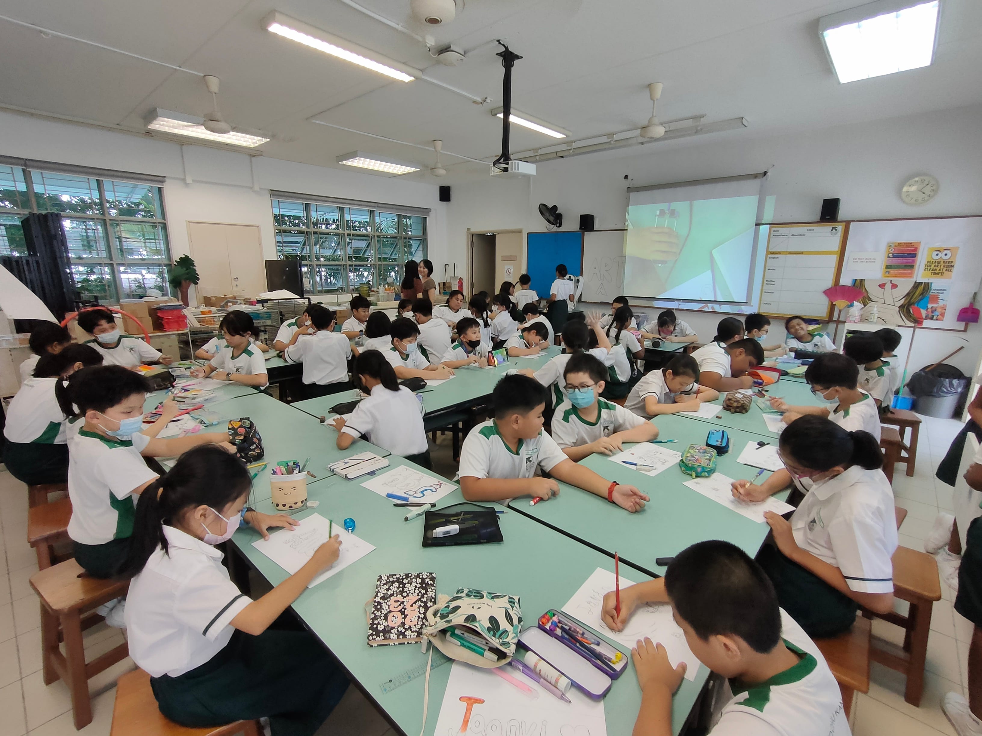 Students in white uniforms and face masks draw at green tables in a bright classroom with a projector screen.